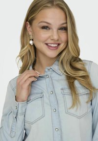 Camicia di jeans blu chiaro con bottoni a pressione, due tasche sul petto e una vestibilità rilassata. Le maniche lunghe sono arrotolate. Orecchini con goccia di perla come accessori.