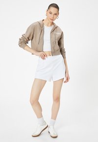 Felpa marrone con cappuccio e zip frontale, decorata con testo bianco, abbinata a pantaloni sportivi bianchi e sneakers bianche con dettagli marroni.