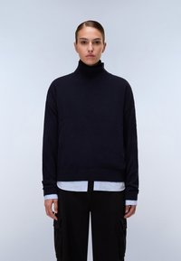 Marineblauer Rollkragenpullover mit strukturiertem Strick, lockerer Passform und gerippten Bündchen, kombiniert mit einem weißen Hemd mit Kragen und schwarzen Cargo-Hosen.