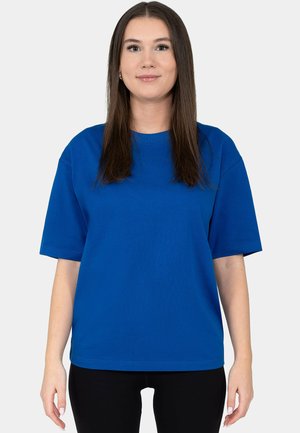 Young woman with straight brown hair wearing a plain blue short-sleeve shirt and black pants, standing against a white background.