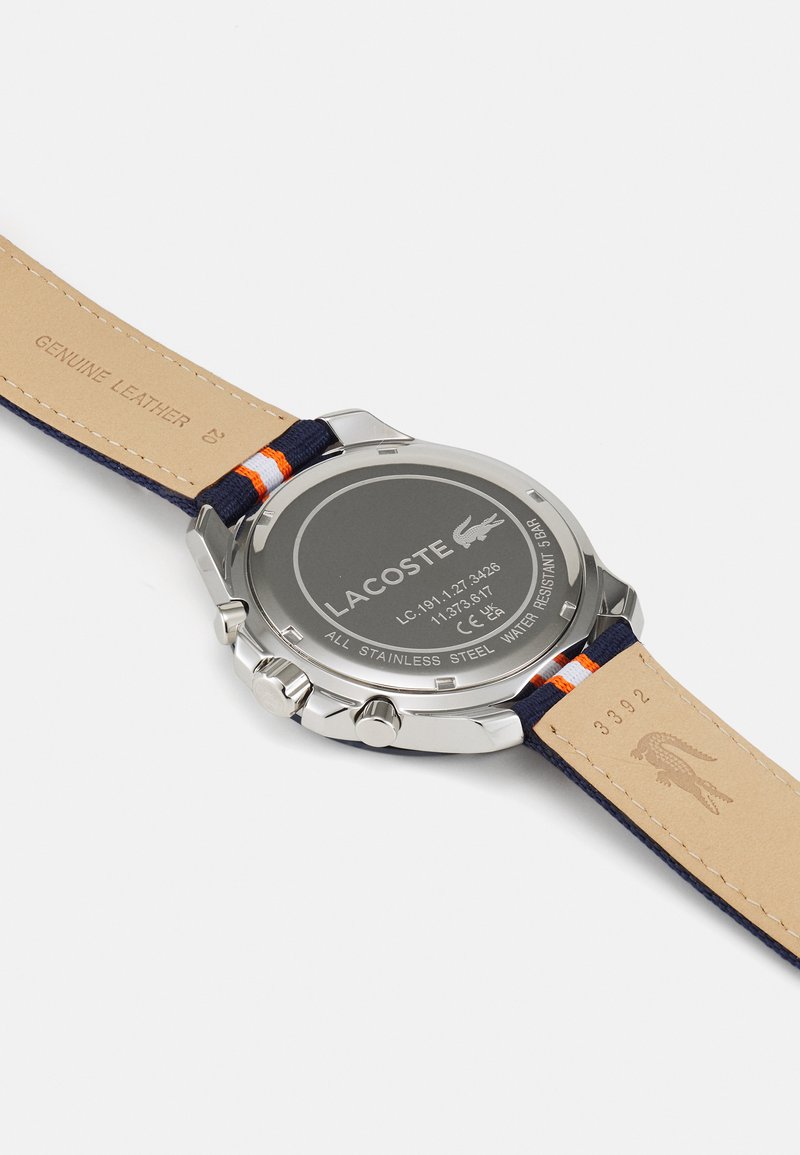 Reloj Lacoste con caja de acero inoxidable, correa de cuero azul marino con acento naranja y logo grabado en la parte posterior.