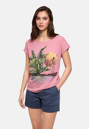 Mujer que lleva una camiseta rosa con un estampado colorido de una persona remando un barco cerca de grandes hojas verdes y un sol naranja, combinada con pantalones cortos azul marino.