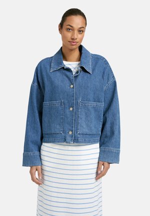 Frau trägt eine blaue Jeansjacke mit großen Fronttaschen über einem weißen Kleid mit hellblauen horizontalen Streifen.