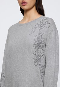Grå sweatshirt med långa ärmar som har svarta blommiga linjeteckningar på vänster axel och ärm. Mjuk, lätt tyg med avslappnad passform.