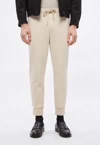 Beżowe spodnie dresowe z elastycznym pasem, sznurkiem i zwężanymi mankietami. Gładka tekstura, minimalne przeszycia i brak widocznych kieszeni.