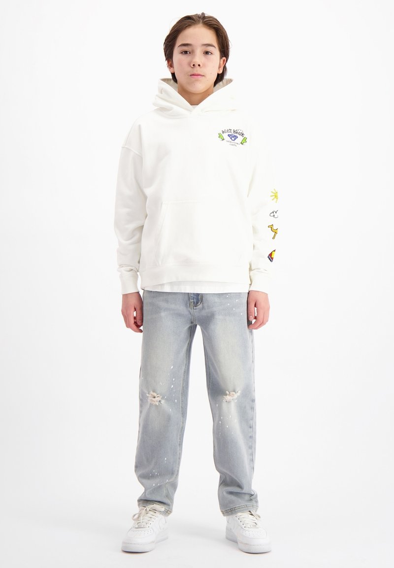 Witte hoodie met een voorzak en grafische accenten, gecombineerd met lichtblauwe ripped jeans met verfspetters. Witte sneakers maken de look compleet.