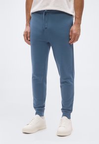 Άνδ ρας που φοράει μπλε παντελόνι αθλητισμού και λευκά αθλητικά παπούτσια, στέκεται μπροστά σε λευκό απλό φόντο.