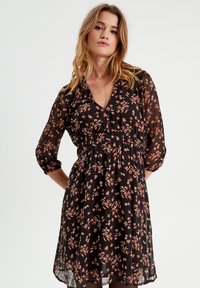 Robe noire à fleurs avec un décolleté en V, des manches trois-quarts, une taille élastique et une texture légère et fluide. Présente de petits motifs floraux roses et blancs.