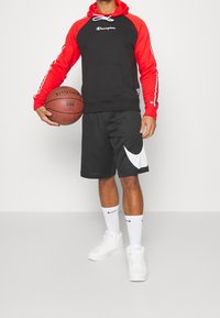 Felpa con cappuccio nera e rossa con dettagli bianchi, pantaloncini neri con logo Nike bianco, scarpe da ginnastica bianche, mentre tiene un pallone da basket.