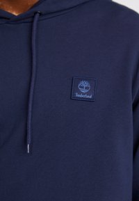 Dunkelblaue Kapuzenjacke aus Baumwolle mit einem kleinen, gestickten Timberland-Logo in Hellblau auf der Brust und einer Kordelzugkapuze.