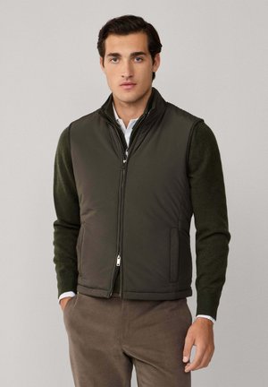Young man wearing olive green zip-up vest over dark green sweater and brown pants, standing against plain light grey background.