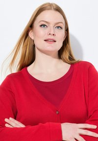 Cardigan rosso con scollatura a V, texture a coste e un solo bottone. Indossato sopra una camicia rossa a strati, con un collo liscio.