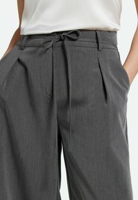 Pantalones grises con una textura suave, que presentan una cintura con un lazo atado y pliegues frontales, exhibidos con las manos en los bolsillos.