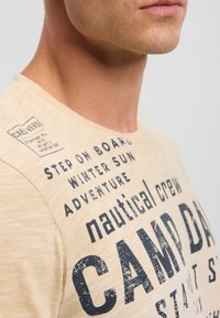Beige t-shirt featuring various black text prints, including "CAMP DAY" and nautical themes, with a soft, textured fabric. Round neckline.