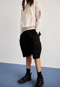 Beżowa bluza z kapturem z pomarańczowym logo, czarne szorty typu cargo i czarne sneakersy. Bluza ma przedni kieszeń oraz kieszeń na rękawie na zamek.