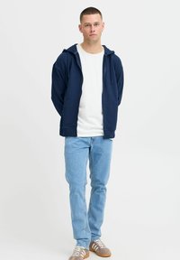 Marineblauwe rits-hoodie met een voorzak, gecombineerd met een wit T-shirt, lichtblauwe spijkerbroek en grijs-bruin sneakers met witte veters.
