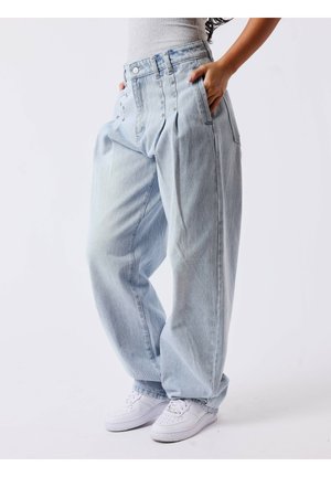 Personne portant un jean bleu clair à jambes larges avec des clous près des poches et des baskets blanches, main dans la poche, sur un fond uni.