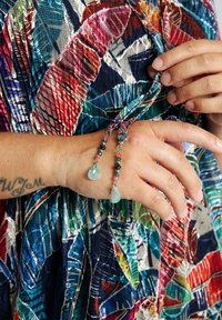 Kädet säätävät värikästä kuviollista kangasta, jonka reunoissa on helmiripustuksia, jotka päättyvät sinisiin vesipisaramaisiin kivenkappaleisiin. Vasemmassa kädessä näkyy tatuointi.