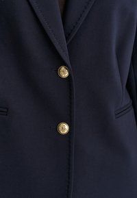 Gros plan sur un blazer bleu marine foncé avec deux boutons décoratifs dorés et des détails de revers cousus, montrant une partie des poches avant.