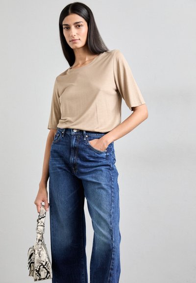 Beige, top met korte mouwen, gecombineerd met hoge taille, wijde blauwe jeans. De persoon houdt een kleine, gestreepte handtas vast met een slangenprint.