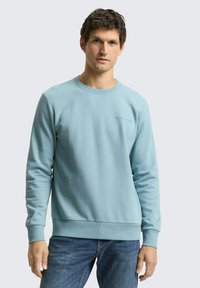 Lichtblauwe sweatshirt met een ronde hals, lange mouwen en geribbelde manchetten. Heeft een subtiel logo op de borst. Gecombineerd met een blauwe spijkerbroek.