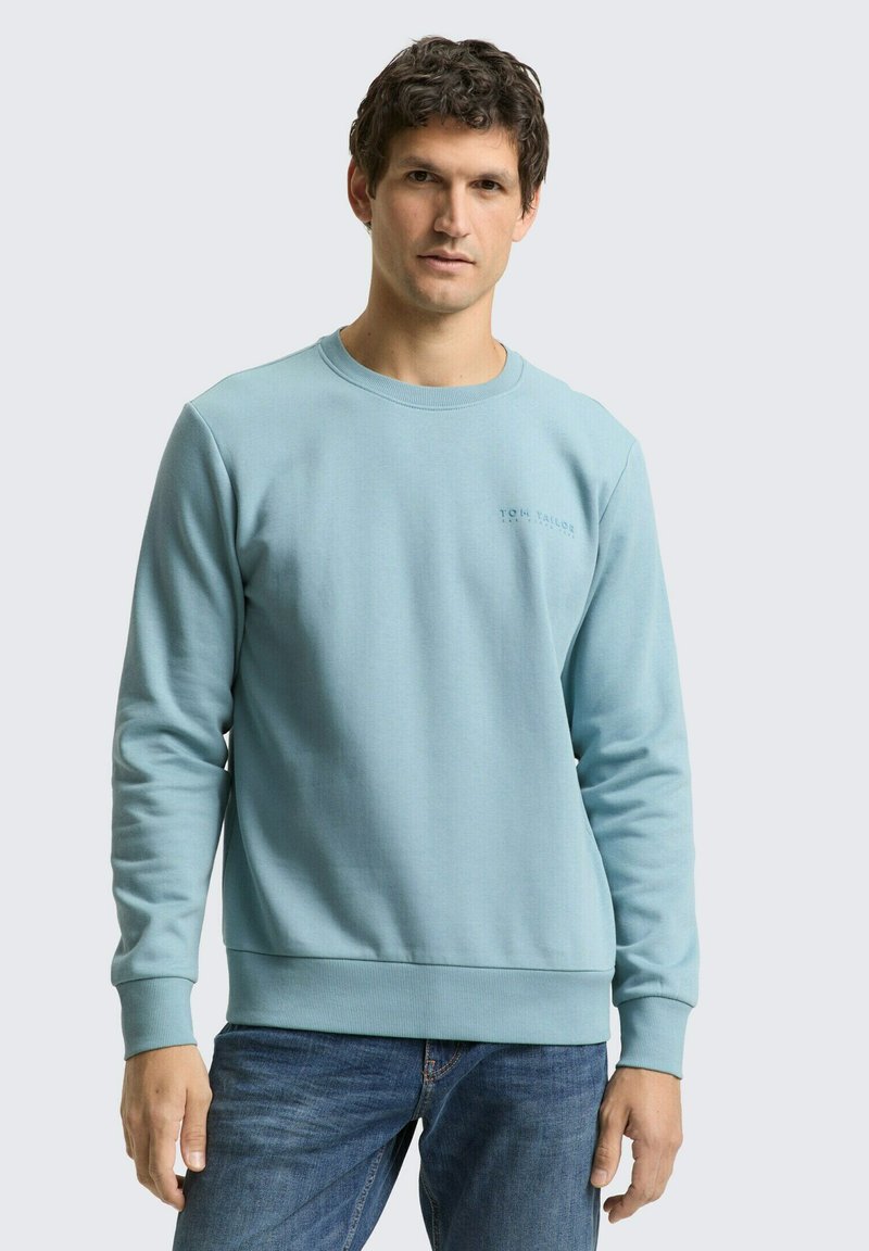 Lichtblauwe sweatshirt met een ronde hals, lange mouwen en geribbelde manchetten. Heeft een subtiel logo op de borst. Gecombineerd met een blauwe spijkerbroek.