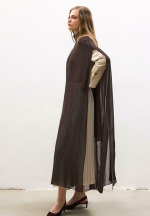 Nő egy hosszú, félig átlátszó fekete pliszírozott ruhában, bézs pliszírozott ruha réteggel alatta, fekete pántos magassarkú cipőben, oldalra fordulva áll.