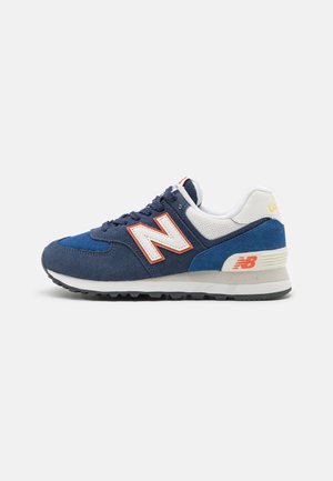 Marineblaue und weiße New Balance Sneaker mit orangefarbenem "N"-Logo, Obermaterial aus Wildleder und Mesh, weißer Zwischensohle und schwarzer Außensohle.