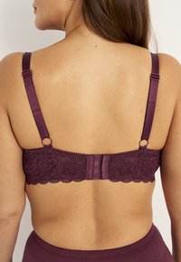 Burgunderfarbene Spitzen-BH mit verstellbaren Satinschulterträgern, versehen mit floralem Muster und Haken- und Ösenverschluss auf der Rückseite. Weicher Stoff.