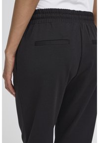 Sorte joggebukser med strukturert stoff, elastisk midjebånd og to baklommer med svarte detaljer.