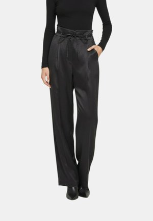 Donna che indossa pantaloni larghi in satin nero a vita alta con cintura paperbag e coulisse, abbinati a scarpe nere con punta appuntita e top nero a maniche lunghe.