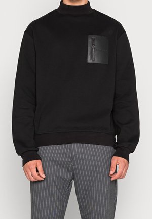 Homme portant un sweat-shirt noir avec une poche poitrine en cuir zippée, associé à un pantalon gris à fines rayures.