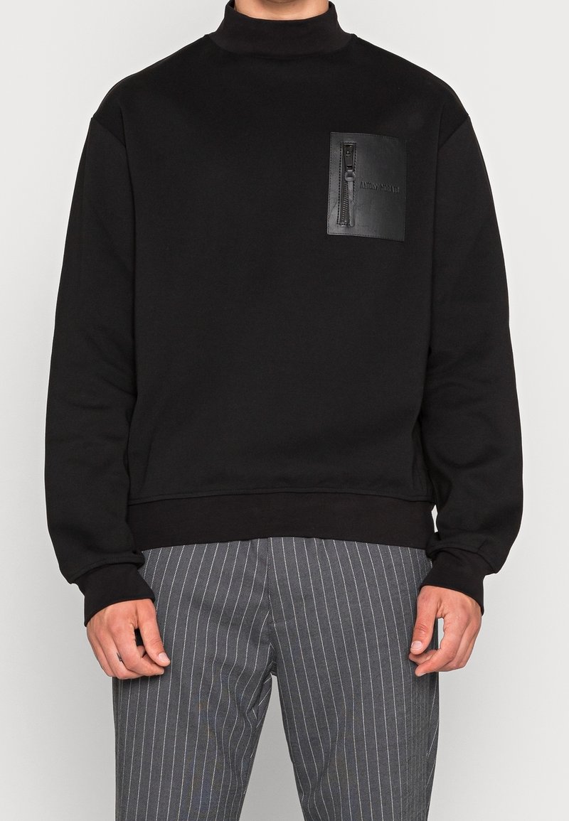 Homme portant un sweat-shirt noir avec une poche poitrine en cuir zippée, associé à un pantalon gris à fines rayures.