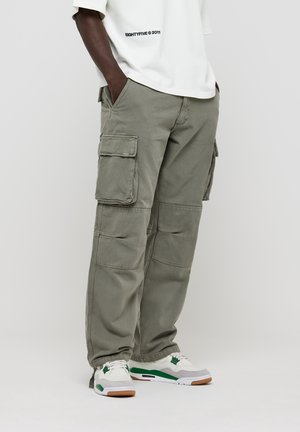 Cargo pants in olive green cotton, featuring large side pockets, a relaxed fit, and visible stitching details. Paired with white and green sneakers.