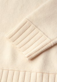 Stickad krämfärgad tröja med detaljer som ribbade ärmar och en mjuk textur, som visar en slät finish och ett subtilt mönster på tyget.