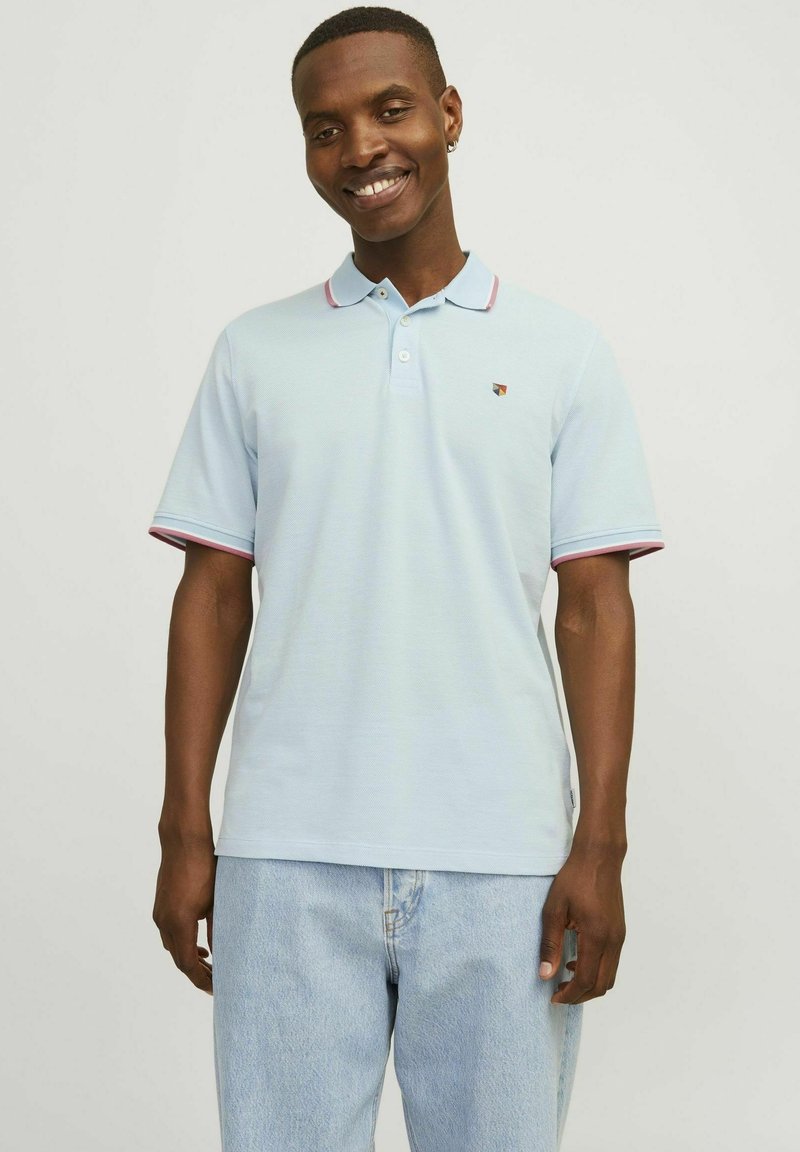 Maglietta polo azzurra con tessuto strutturato, rifiniture rosa a contrasto su maniche e colletto, e un piccolo logo sul lato sinistro del petto. Abbinata a un paio di jeans chiari.