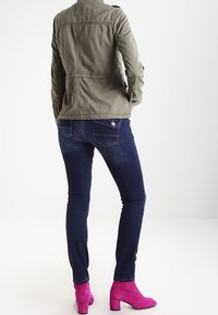 Olivengrøn jakke med tætsiddende design, kombineret med mørkeblå skinny jeans og lyserøde ankelstøvler. Enkel syning og knappedetalje er synlige.