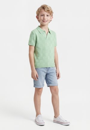 Ξανθός αγόρι που στέκεται, φοράει ανοιχτό πράσινο κοντομάνικο πουκάμισο με γιακά, ανοιχτό μπλε τζιν σορτς με γυρισμένα μπατζάκια και λευκά αθλητικά παπούτσια.