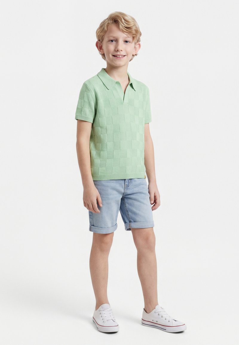 Ξανθός αγόρι που στέκεται, φοράει ανοιχτό πράσινο κοντομάνικο πουκάμισο με γιακά, ανοιχτό μπλε τζιν σορτς με γυρισμένα μπατζάκια και λευκά αθλητικά παπούτσια.