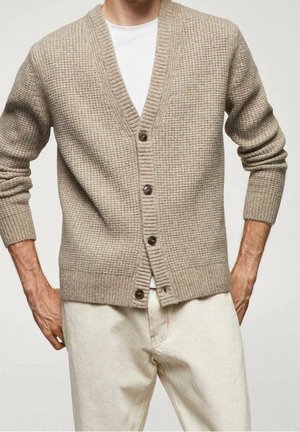 Homme portant un cardigan beige texturé boutonné sur une chemise blanche, associé à un pantalon clair, les mains partiellement dans les poches.