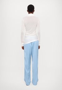 Langärmlige, durchsichtige weiße Bluse, kombiniert mit hellblauen, weit geschnittenen Hosen. Das Ensemble zeichnet sich durch ein drapiertes Design und glatte Texturen aus.