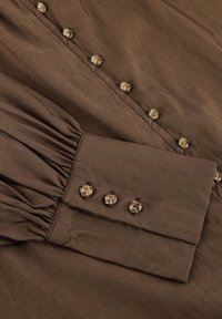 Bruine stof met een gestructureerde afwerking, voorzien van een knoopsluiting versierd met bloemenvormige gouden knopen en gathered cuffs.