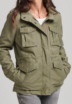 Frau trägt eine olivgrüne Utility-Jacke mit mehreren Klappentaschen, darunter ein weißes Oberteil und eine silberne Anhänger-Halskette.