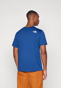 Ανδρικό μπλε κοντομάνικο μπλουζάκι, με στρογγυλή λαιμόκοψη, φτιαγμένο από απαλή ύφανση, διαθέτει ένα μικρό λευκό λογότυπο στην πάνω πλάτη, συνδυασμένο με πορτοκαλί παντελόνι.