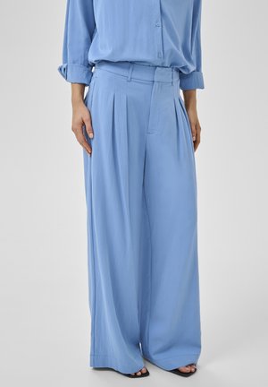 Világoskék, széles szárú nadrág elöl pliszírozva, hozzá illő hosszú ujjú gombos inggel és fekete, nyitott orrú szandállal viselve.