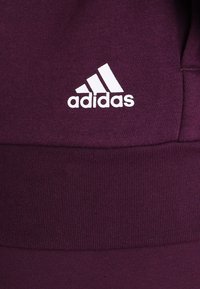 Sudadera morada de Adidas con una tela texturizada, que presenta un logo blanco. El diseño incluye un dobladillo acanalado para un detalle adicional.