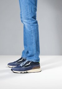 Pánské tenisky v námořnicky modré semišové a síťované kombinaci, s bílou stélkou, podrážkou ve tvaru cikcak a zeleným akcentem na patě. Nošeny s modrými džíny.