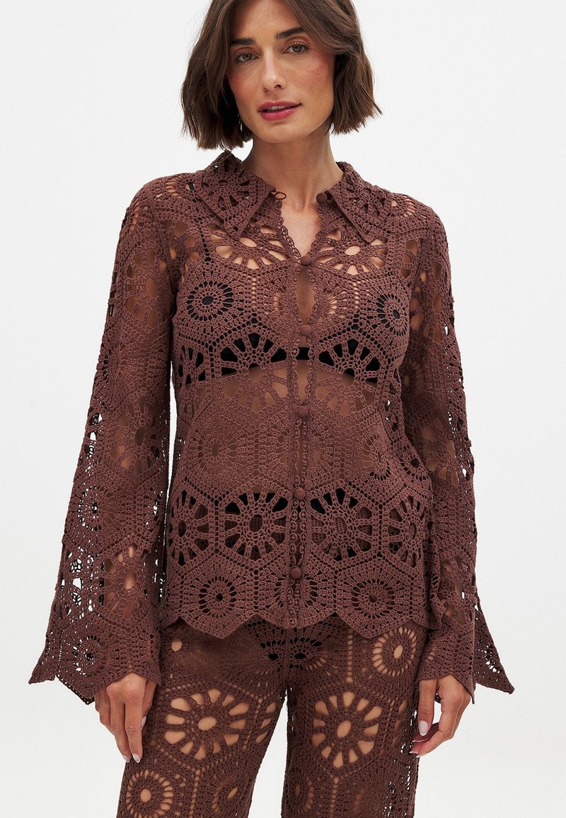 Crochet blouse in dark brown, featuring intricate floral patterns, a collared neckline, and flared sleeves. Material appears lightweight and textured.