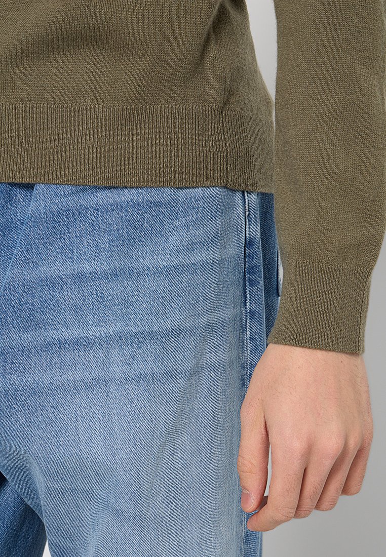 Olivgrön stickad tröja med ribbade muddar, ihopparad med ljusblå jeans i denim med en blekt finish. Närbild av arm och midja.