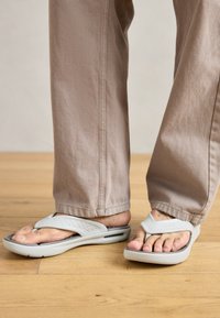 Jasnoszare klapki z teksturowanym paskiem i perforowanymi detalami, spoczywające na drewnianej podłodze, z widocznymi częściowo beżowymi spodniami.